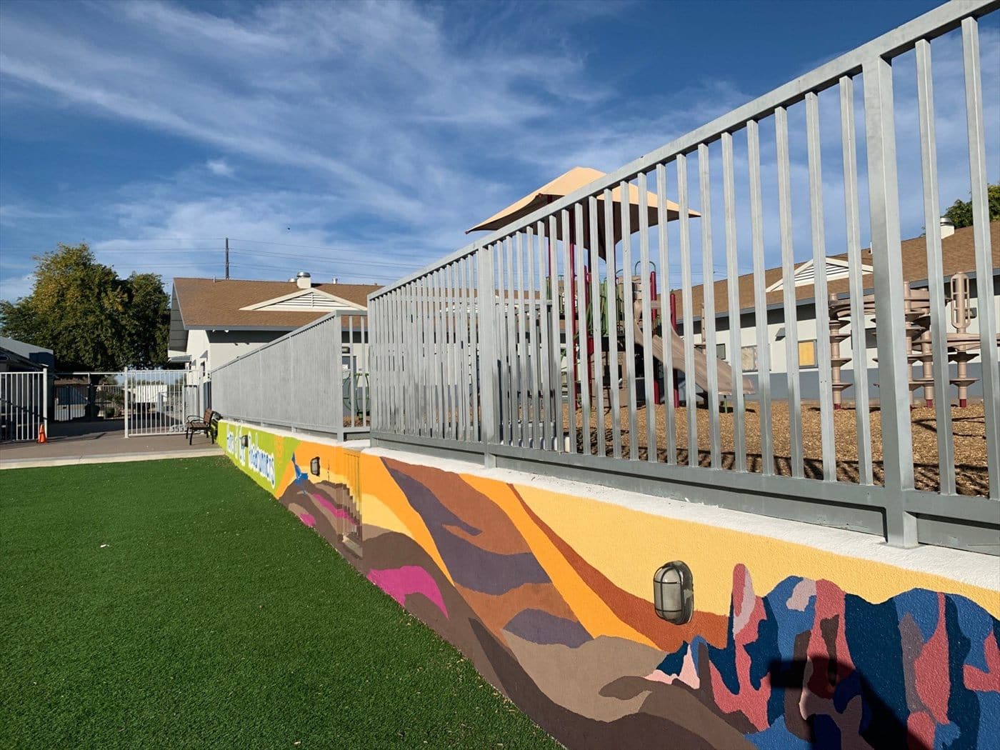 Repainted gray fence atop a colorful mural retaining wall beside artificial turf.