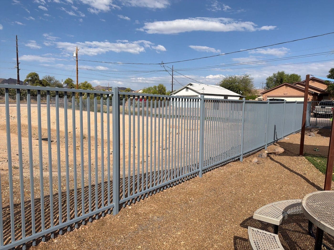 Repainted gray picket-top perimeter fence bordering a dirt lot with houses beyond.