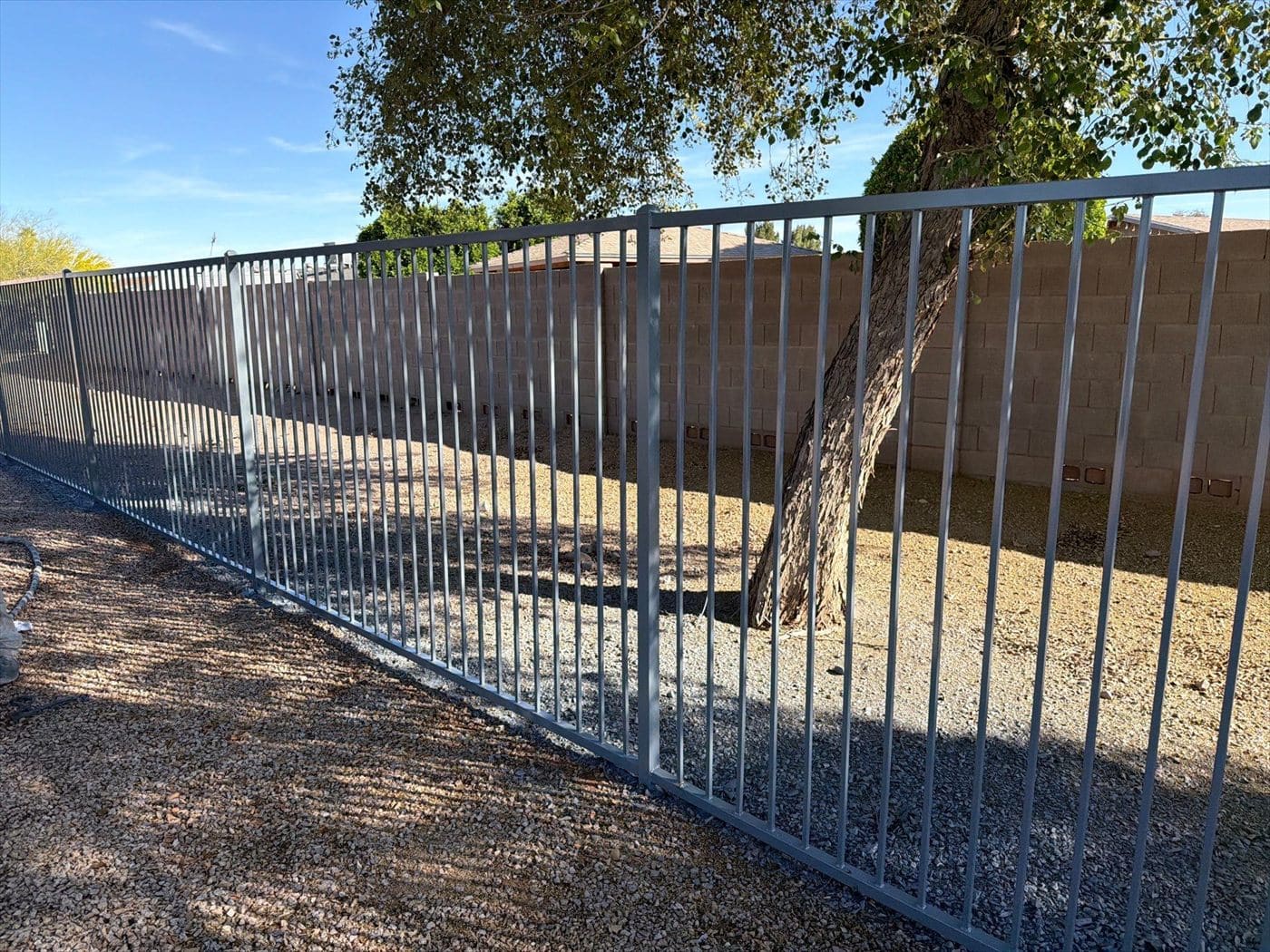 Gray perimeter metal fence surrounding a gravel yard with a tree and block wall behind.