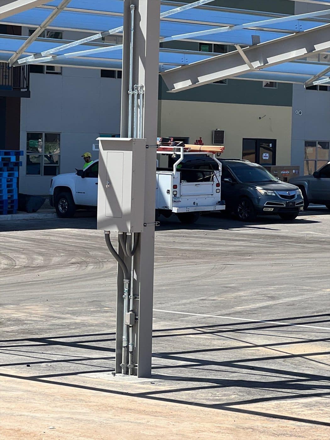 A gray painted electrical box and conduit on a carport column with service trucks in the lot.