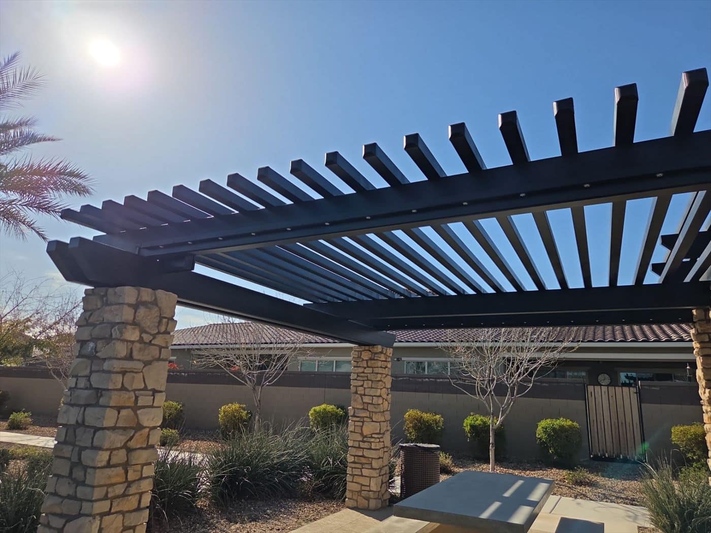 Black louvered pergola on stone columns beside a community building.
