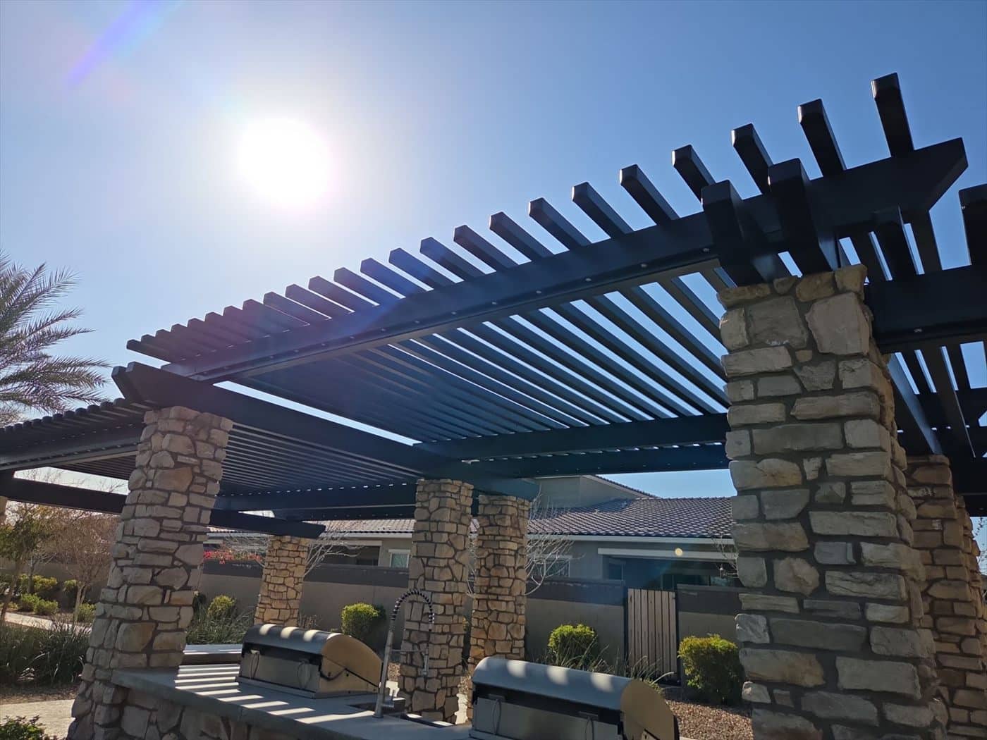 Sun over a black metal pergola with stone columns and BBQ stations.