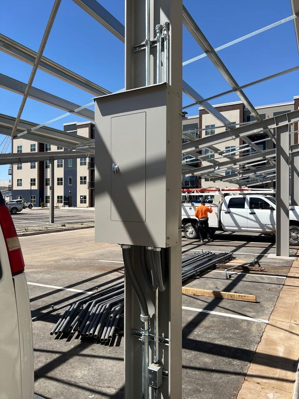 Close-up of a gray painted electrical box and conduit on a solar carport column with work trucks behind.