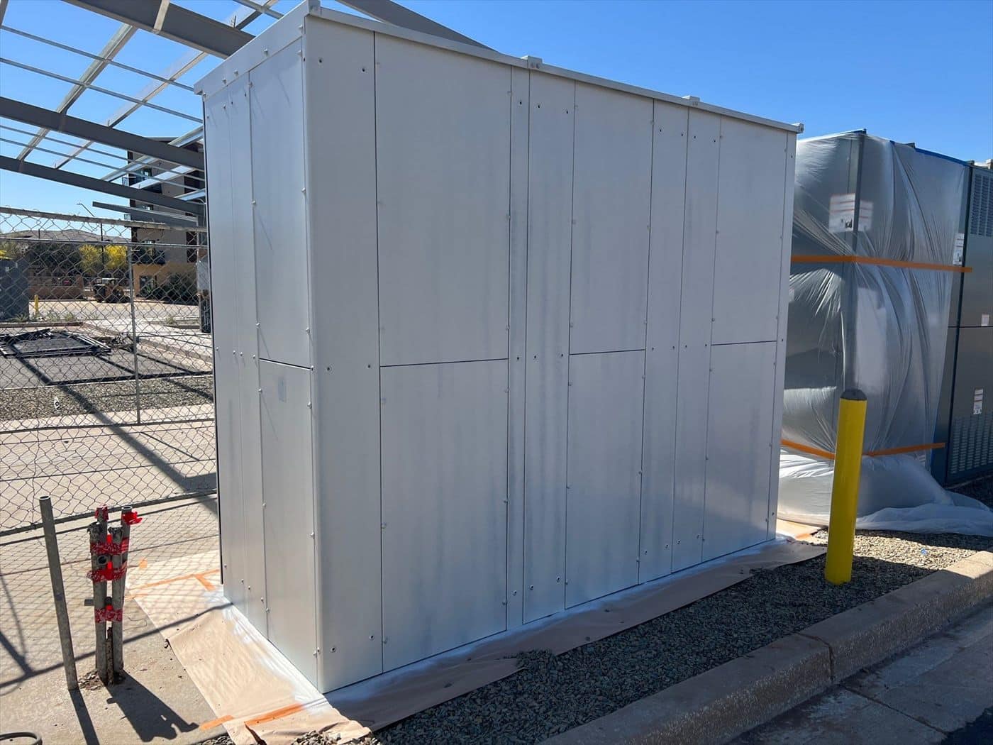 A freshly painted white metal equipment enclosure with a plastic-wrapped unit beside it.